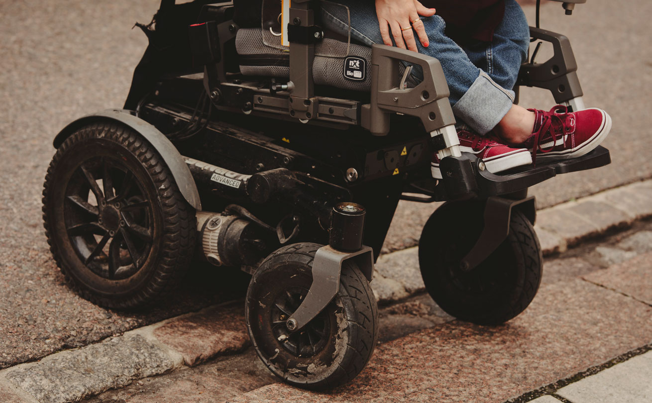 Lähikuva sähköpyörätuolin pyörästä, kuvassa näkyvät myös Anni Kyröläisen jalat.
